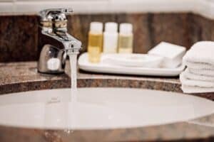 A bathroom sink with running water, a faucet, stacked white towels, and toiletries in small bottles on a tray adorn the countertop—showcasing the results of a recent hotel plumbing upgrade.