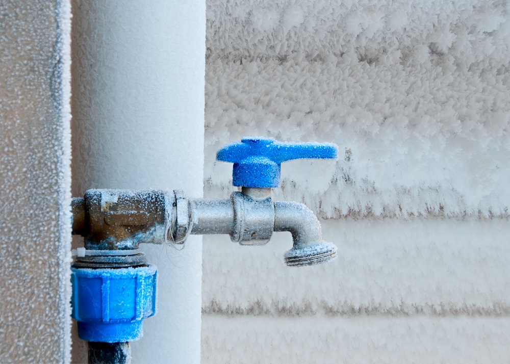 A metal outdoor water faucet with a blue handle is covered in frost, indicating freezing temperatures. Frost is visible on the adjacent wall and pipe—highlighting the need for pipe insulation in Tacoma’s cold climate.