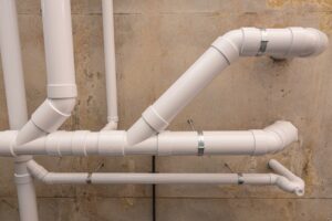 Two white vent pipes in bathroom plumbing run vertically up a tiled wall, connecting to a valve or fixture near the ceiling at the corner of the room.