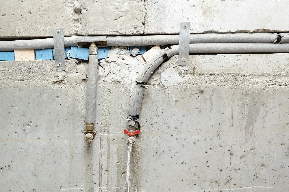Exposed pipes and cables run along a damaged concrete wall, some wrapped in Pipe Insulation Tacoma materials and secured by metal brackets. A red tape is visible on one section of pipe.