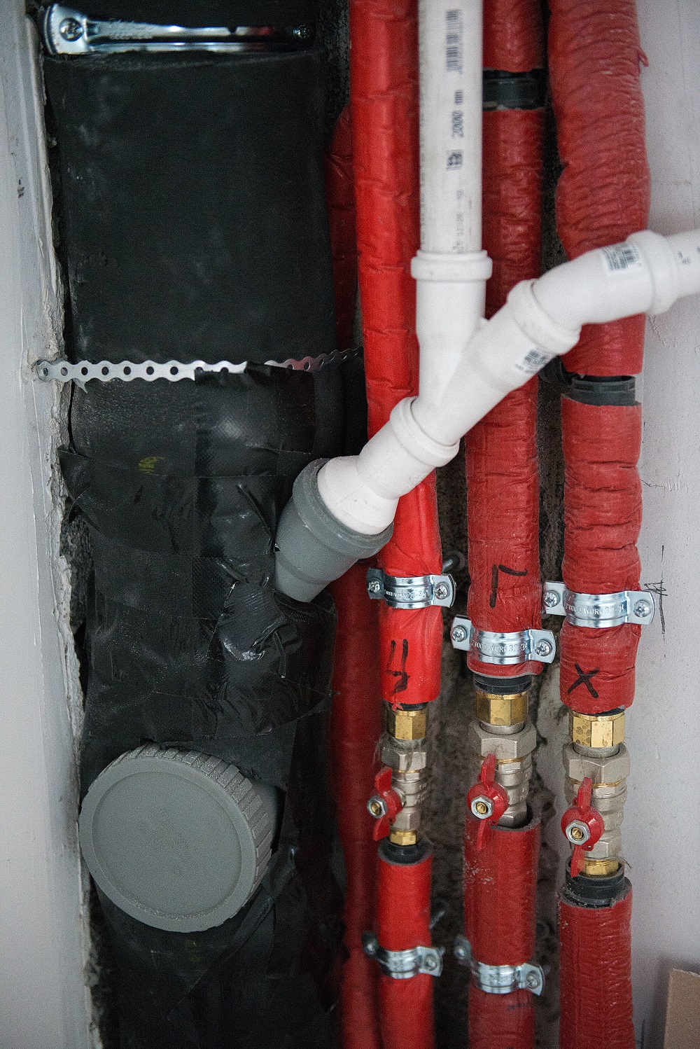 Plumbing pipes with red insulation and metal fittings, including a black pipe with a grey cap and a white plastic connector, mounted against a wall—showcasing quality pipe insulation Tacoma homeowners trust.