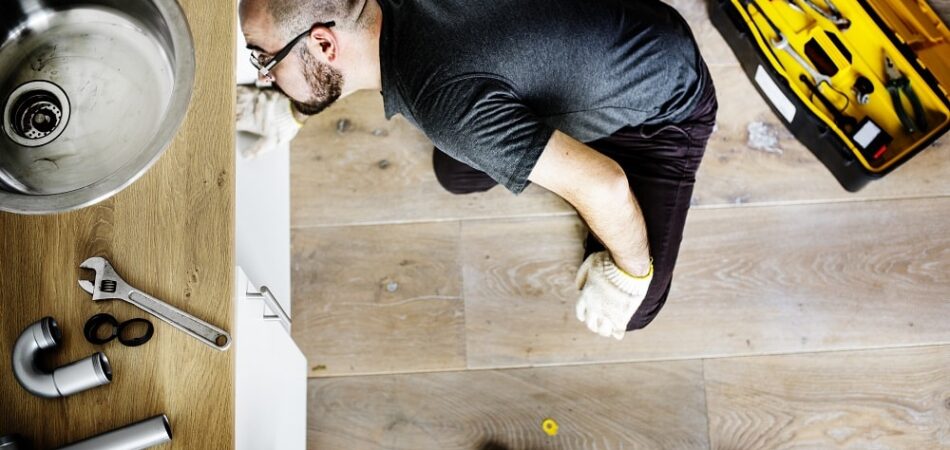 A person kneels on a wooden floor fixing plumbing under a sink, surrounded by pipes and tools, with an open toolbox nearby—much like professional plumbers in Olympia at work.