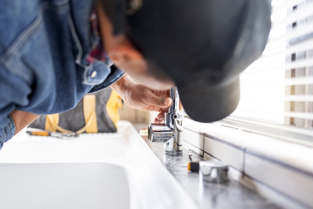 A person wearing a cap and denim shirt repairs a kitchen faucet next to a sink, with tools and a tool bag visible nearby—showcasing reliable Plumbing Solutions Gig Harbor in action.