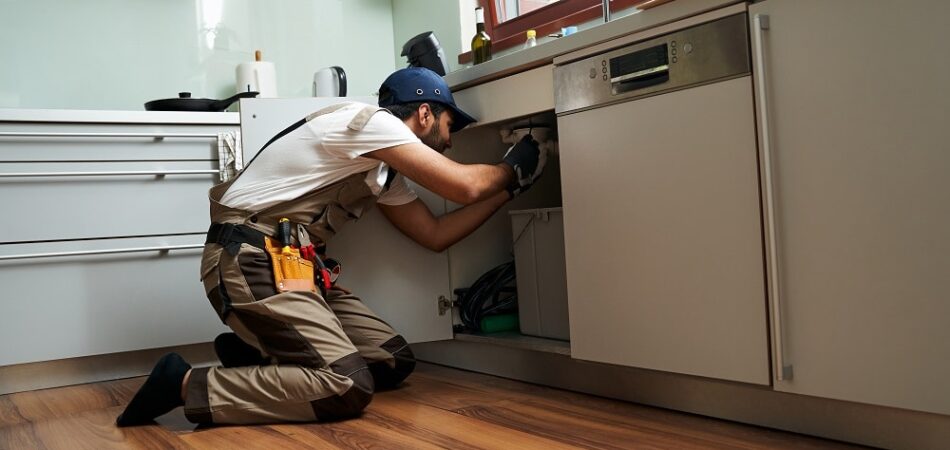 A person wearing work clothes and gloves is kneeling on the floor, repairing or inspecting the plumbing under a kitchen sink—demonstrating expert Plumbing Services in Graham.