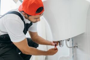 A plumber offering Plumbing Services in Spanaway, wearing a red cap and overalls, uses a screwdriver to fix plumbing beneath a white bathroom sink. A plumber offering Plumbing Services in Spanaway, wearing a red cap and overalls, uses a screwdriver to fix plumbing beneath a white bathroom sink.