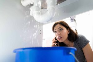A woman inspects a leaking pipe above a blue bucket, capturing water cascading down into the bucket