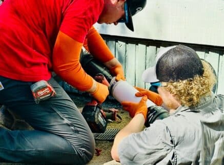Two workers wearing gloves and caps use tools to connect or repair a white PVC pipe outdoors next to a wooden fence, ensuring the integrity of private sewer systems.