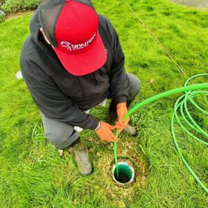 Person wearing orange gloves and a red hat feeds a green hose into an outdoor ground pipe on a grassy lawn.