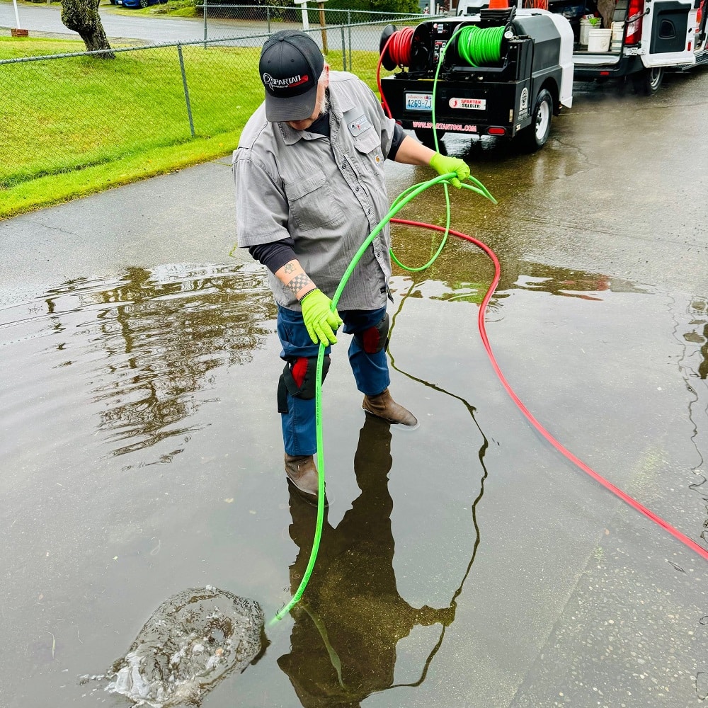A worker wearing gloves and boots stands in a flooded area, holding green and red hoses, with water service equipment, vehicles, and Private Sewer Systems visible in the background.