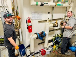 Two workers in safety gear stand by industrial pipes and valves, holding tools and parts near a private sewer systems maintenance area, with a fire extinguisher and various equipment in view.