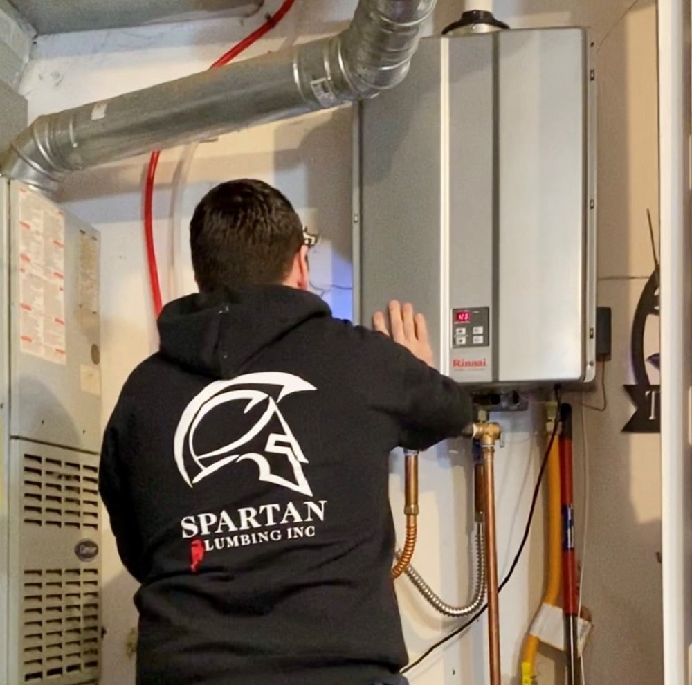 A plumber wearing a Spartan Plumbing Inc hoodie inspects a wall-mounted tankless water heater in a utility room.