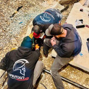 Three people in "SPARTAN" hoodies and caps work together on a rocky ground, appearing to fix or inspect a pipe near a concrete slab—tools and piping materials suggest they're working on Private Sewer Systems.