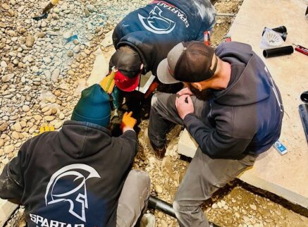 Three people in "SPARTAN" hoodies and caps work together on a rocky ground, appearing to fix or inspect a pipe near a concrete slab—tools and piping materials suggest they're working on Private Sewer Systems.