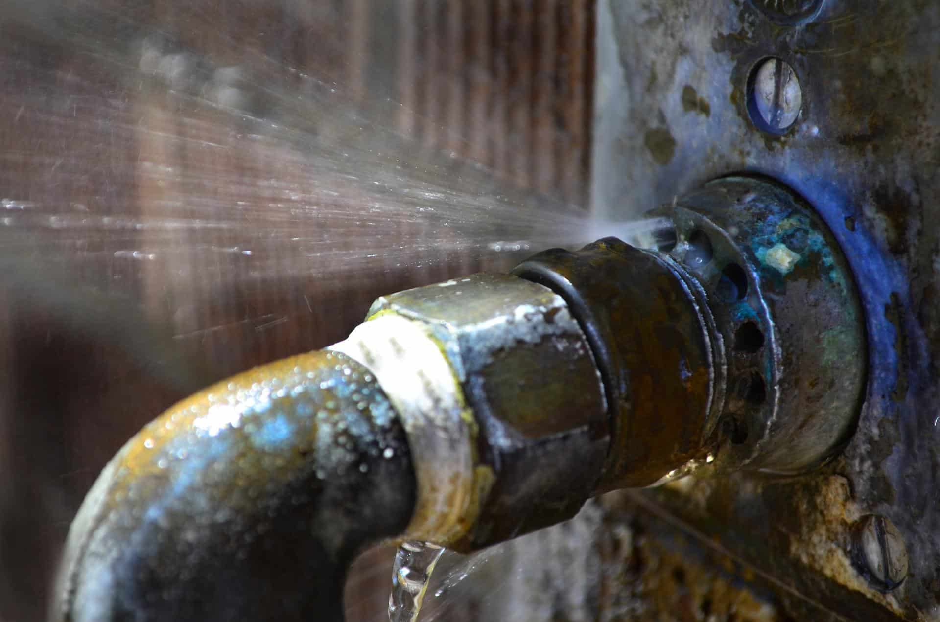Close-up of a leaking pipe joint, with water spraying and droplets visible against a rustic background. Signs of corrosion are present.