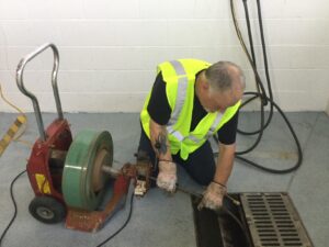 Man in a neon safety vest using drain cleaning equipment. He kneels beside a floor drain, focused and concentrated, with gloves for protection.