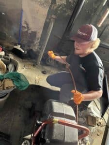 Benji from Spartan Plumbing wearing orange gloves and a cap is handling a copper pipe next to a large machine in a cluttered, dimly lit room, showcasing residential plumbing in action.