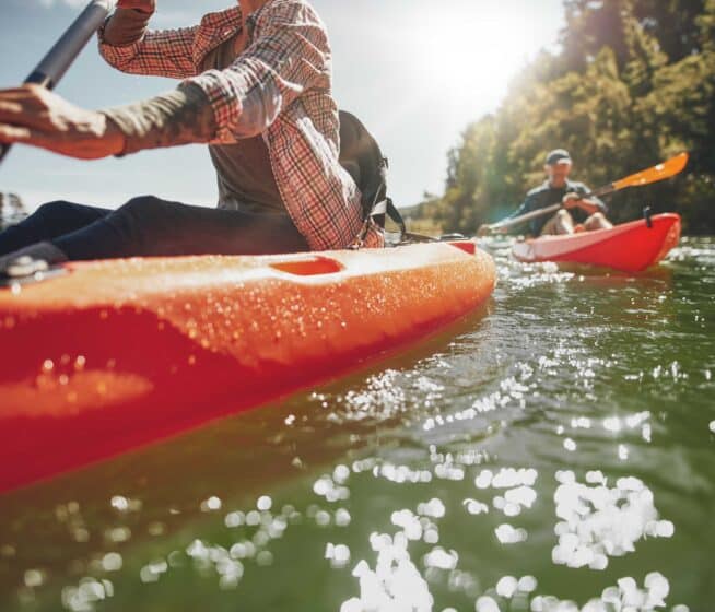 Two people paddle bright red kayaks on a sunlit river, with trees visible in the background and water reflecting sunlight.