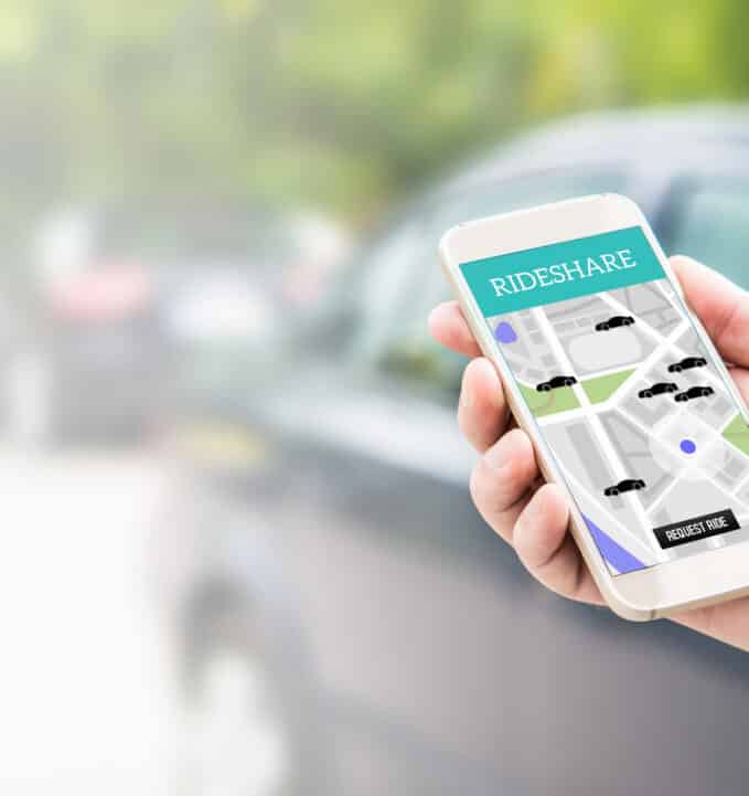 A person holds a smartphone displaying a rideshare app with a map and car icons, with a blurred car in the background.