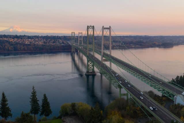 Aerial view of a long suspension bridge crossing a body of water at sunset, with cars on the roadway and a distant snow-capped mountain in the background.