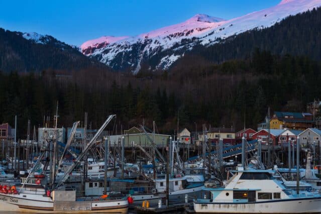 Fishing boats docked in a harbor with snow-capped mountains and colorful buildings in the background at sunrise or sunset.