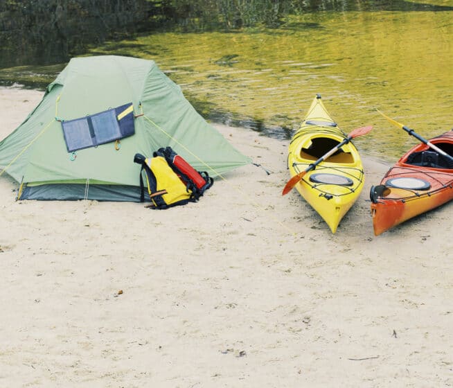 A green tent with a solar panel and backpacks beside it stands on sandy ground near a river, next to a yellow and an orange kayak with paddles.