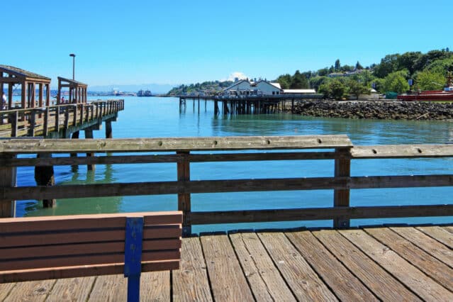 Wooden pier with bench overlooking calm water, docks, and buildings in the distance under a clear blue sky.